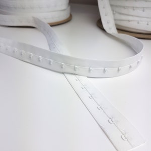 May include: White hook and eye tape, suitable for various sewing applications. The tape is made of a woven fabric and has evenly spaced metal hooks and eyes. Two spools of the tape are visible in the background.