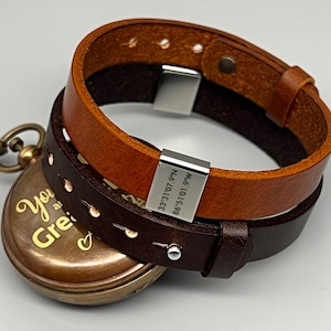 May include: A brown leather bracelet with a silver clasp and a silver tag engraved with coordinates. The bracelet is wrapped around a pocket watch with a gold-colored face and the inscription "You are great" on it.