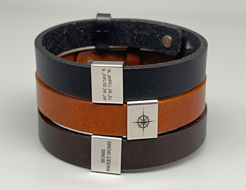May include: Three leather bracelets with silver charms. The top bracelet is black leather with a silver charm that reads "40&deg; 44' 55-7412" N 73&deg; 59' 7.5408" W.". The middle bracelet is brown leather with a silver compass charm. The bottom bracelet is dark brown leather with a silver charm that reads "HOME SWEET HOME".