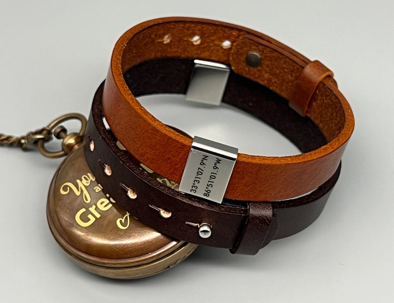 May include: A brown leather bracelet with a silver clasp and a silver tag engraved with coordinates. The bracelet is wrapped around a pocket watch with a gold-colored face and the inscription "You are great" on it.