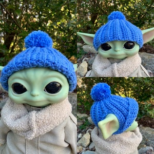 May include: A blue knitted hat with a pom pom on top. The hat is crocheted and has a ribbed texture. The hat is worn by a toy figure with large eyes and green skin.
