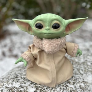Op de afbeelding: Een klein groen pluche speelgoedje met grote ogen en een beige jasje staat op een besneeuwde rots. Het speelgoed heeft een zachte, harige kraag en draagt een beige jasje.