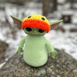 May include: A green plush toy wearing a yellow and orange crocheted hat with black buttons. The toy is standing on a rock.