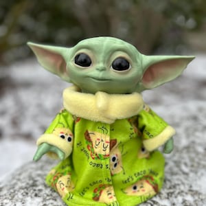 May include: A green plush toy with large eyes wearing a green and yellow robe with a taco print and the text "Let's taco 'bout it".