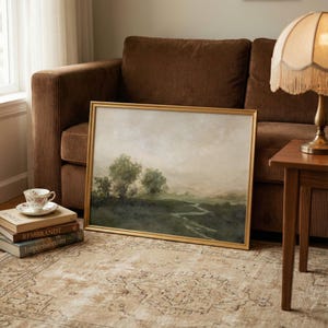 May include: A framed landscape painting with a gold-colored frame, depicting a serene scene with trees and a winding path. The painting is leaning against a brown corduroy sofa, with a teacup and books nearby.
