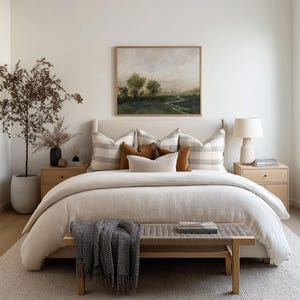 May include: A bedroom with neutral tones features a bed with a beige duvet and decorative pillows. A landscape painting hangs above the bed. A wooden bench with a blanket sits at the foot of the bed. A rug and two nightstands complete the room.