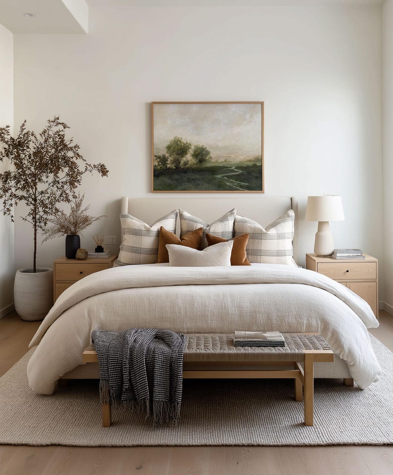 May include: A bedroom with neutral tones features a bed with a beige duvet and decorative pillows. A landscape painting hangs above the bed. A wooden bench with a blanket sits at the foot of the bed. A rug and two nightstands complete the room.