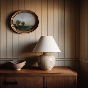 May include: A cream-colored ceramic lamp with a white lampshade sits on a wooden cabinet. A small, oval painting in a wooden frame hangs on the wall above the lamp. A small, beige bowl is on the cabinet to the left of the lamp. The wall is paneled.
