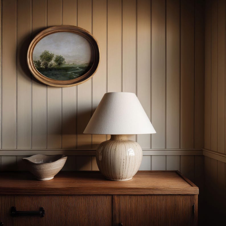 May include: A cream-colored ceramic lamp with a white lampshade sits on a wooden cabinet. A small, oval painting in a wooden frame hangs on the wall above the lamp. A small, beige bowl is on the cabinet to the left of the lamp. The wall is paneled.