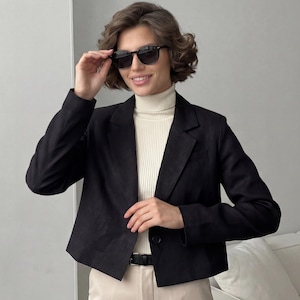 Op de afbeelding: Een zwarte blazer met een enkele knoopsluiting, gedragen over een crèmekleurige coltrui. De blazer wordt gecombineerd met lichtgekleurde broeken en een zwarte riem. De persoon draagt een zwarte zonnebril.