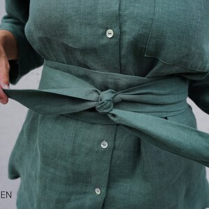 May include: A green linen shirt with a matching green linen belt tied around the waist. The shirt has a button-up front and long sleeves.