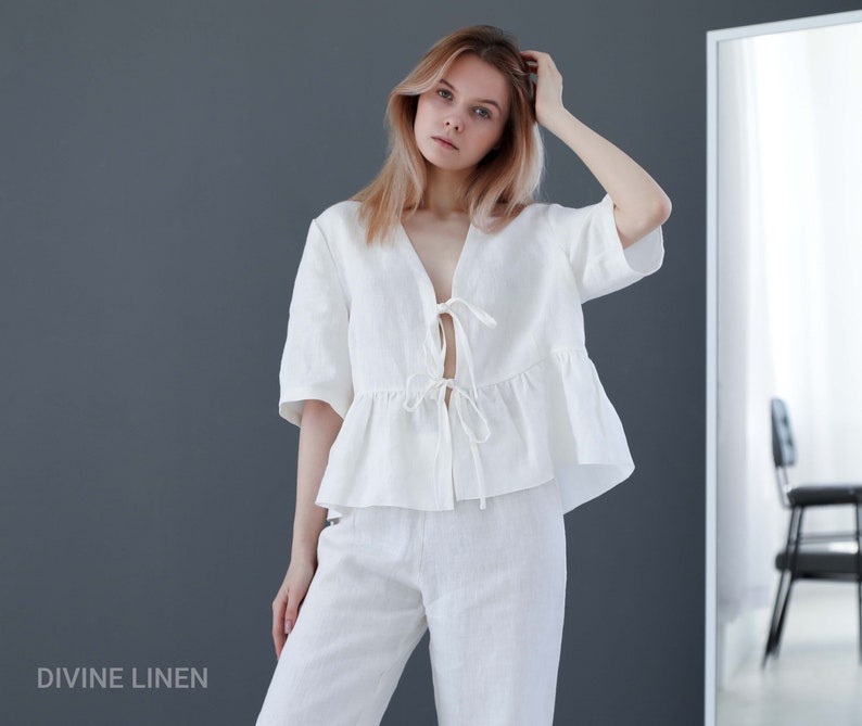 Puede incluir: Una blusa blanca de lino con un lazo en el escote y un dobladillo con volantes. La blusa se combina con pantalones blancos de lino. La modelo lleva el conjunto y est&aacute; de pie frente a un espejo.