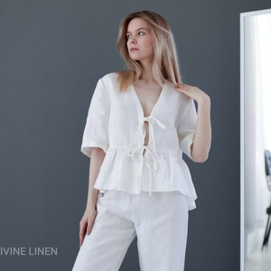 Puede incluir: Una blusa blanca de lino con cuello en V y un lazo en la parte delantera. La blusa tiene un dobladillo con volantes y se lleva con pantalones blancos de lino a juego. La modelo est&aacute; de pie frente a un espejo.