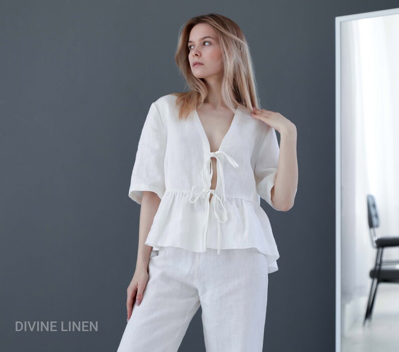 Puede incluir: Una blusa blanca de lino con cuello en V y un lazo en la parte delantera. La blusa tiene un dobladillo con volantes y se lleva con pantalones blancos de lino a juego. La modelo est&aacute; de pie frente a un espejo.