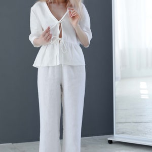 Puede incluir: Un pantal&oacute;n de lino blanco con una blusa de nudo. El pantal&oacute;n es de pierna ancha y la blusa tiene un dobladillo con volantes. La modelo lleva sandalias blancas.