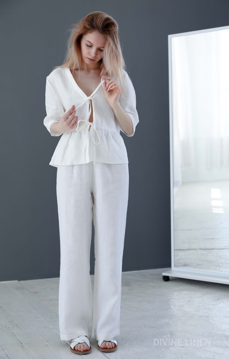 Puede incluir: Un pantal&oacute;n de lino blanco con una blusa de nudo. El pantal&oacute;n es de pierna ancha y la blusa tiene un dobladillo con volantes. La modelo lleva sandalias blancas.