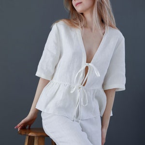 Puede incluir: Una blusa blanca de lino con un lazo en la parte delantera y pantalones a juego. La blusa tiene un dobladillo con volantes y un escote en V profundo. La modelo lleva el atuendo y est&aacute; sentada en un taburete de madera. El texto "DIVINE LINEN" es visible en la esquina inferior derecha de la imagen.