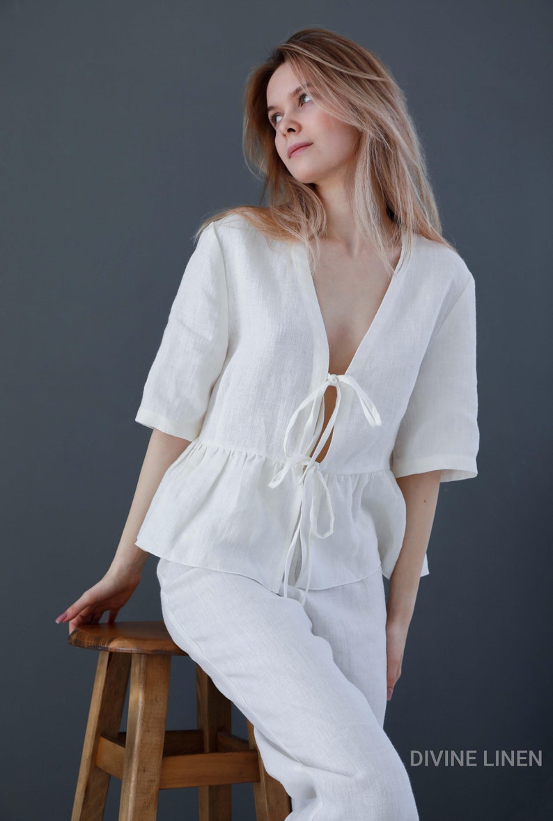 Puede incluir: Una blusa blanca de lino con un lazo en la parte delantera y pantalones a juego. La blusa tiene un dobladillo con volantes y un escote en V profundo. La modelo lleva el atuendo y est&aacute; sentada en un taburete de madera. El texto "DIVINE LINEN" es visible en la esquina inferior derecha de la imagen.