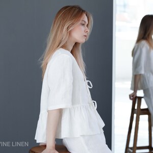 Puede incluir: Una blusa blanca de lino con un dobladillo con volantes y un lazo en la parte delantera. La blusa se lleva con pantalones blancos de lino. La modelo est&aacute; sentada en un taburete de madera.