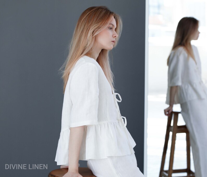 Puede incluir: Una blusa blanca de lino con un dobladillo con volantes y un lazo en la parte delantera. La blusa se lleva con pantalones blancos de lino. La modelo est&aacute; sentada en un taburete de madera.