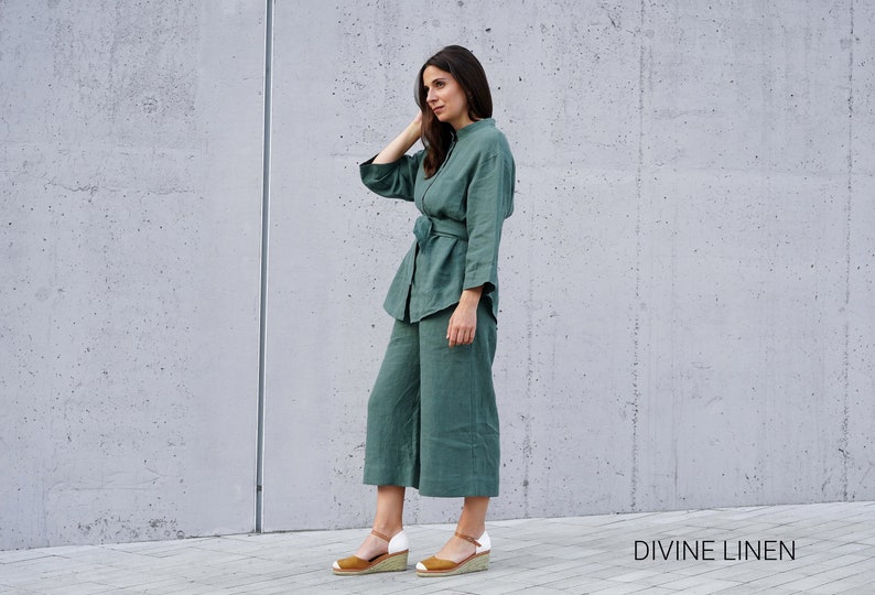 May include: A woman wearing a green linen pantsuit with a matching belt. The pants are cropped and the shirt has a relaxed fit. The woman is standing in front of a concrete wall. The text "DIVINE LINEN" is visible at the bottom of the image.