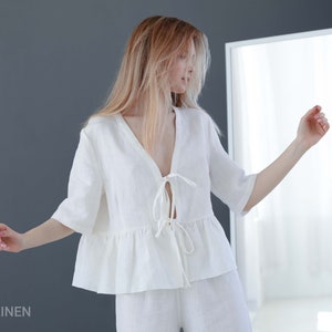 Puede incluir: Una blusa blanca de lino con cuello en V y un lazo en la parte delantera. La blusa tiene un dobladillo con volantes y mangas cortas. La modelo lleva la blusa con pantalones blancos de lino.