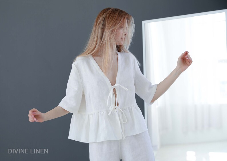 Puede incluir: Una blusa blanca de lino con cuello en V y un lazo en la parte delantera. La blusa tiene un dobladillo con volantes y mangas cortas. La modelo lleva la blusa con pantalones blancos de lino.