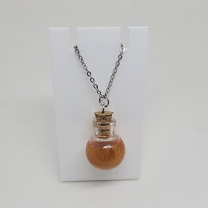 May include: A silver chain necklace with a small glass bottle pendant. The bottle has a cork stopper and is filled with a golden liquid. The bottle is round and has a wooden-like texture.