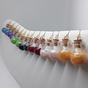 Puede incluir: Una fila de 12 ganchos de pendientes dorados con pequeñas botellas de vidrio llenas de purpurina. Las botellas están llenas de purpurina de diferentes colores, incluyendo azul, morado, verde, negro, rojo, rosa y dorado.