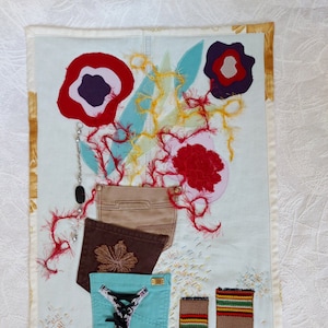May include: A blue and white fabric wall hanging with a flower design. The flowers are made of red, white and purple fabric. The wall hanging also features a brown fabric pocket with a lace detail.