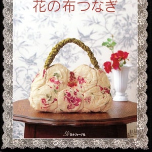 May include: A white fabric handbag with a floral print and a green handle. The bag is sitting on a wooden table with a white vase and red flowers in the background. The text on the cover reads "岡本洋子の おしゃれなパッチワーク 花の布つなぎ".