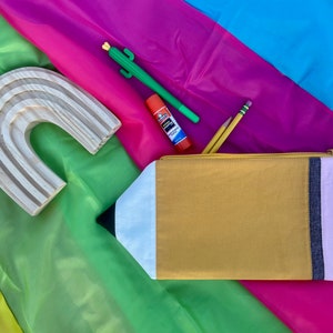Puede incluir: Una bolsa de cremallera amarilla con forma de l&aacute;piz con ribete blanco y rosa, un arco&iacute;ris de madera, un cactus verde, un pegamento en barra y dos l&aacute;pices sobre un fondo de tela multicolor.