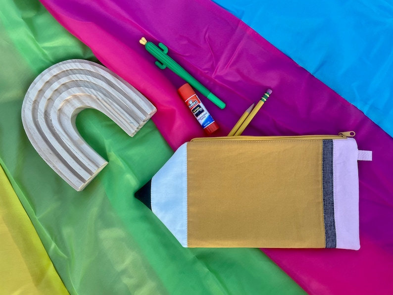 Puede incluir: Una bolsa de cremallera amarilla con forma de l&aacute;piz con ribete blanco y rosa, un arco&iacute;ris de madera, un cactus verde, un pegamento en barra y dos l&aacute;pices sobre un fondo de tela multicolor.