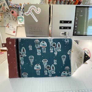 Puede incluir: Una bolsa azul con cremallera con fondo blanco y un patr&oacute;n de medusas. La bolsa tiene un panel de tela marr&oacute;n y negro en el lado izquierdo y un panel de tela dorado en el lado derecho. La bolsa est&aacute; sobre una superficie blanca.