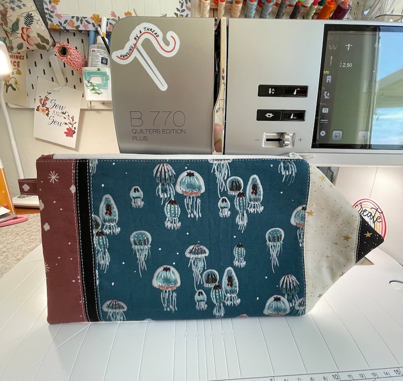 Puede incluir: Una bolsa azul con cremallera con fondo blanco y un patr&oacute;n de medusas. La bolsa tiene un panel de tela marr&oacute;n y negro en el lado izquierdo y un panel de tela dorado en el lado derecho. La bolsa est&aacute; sobre una superficie blanca.