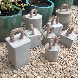 May include: A collection of eight concrete doorstops with rope handles. The doorstops are various sizes and shapes, and are arranged on a bed of small, light brown stones.