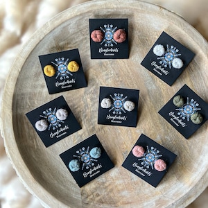 Könnte beinhalten: Sortiment handgefertigter Makrame-Ohrringe in verschiedenen Farben, darunter Braun, Gelb, Beige, Grün und Rosa. Jedes Paar Ohrringe wird auf einer schwarzen Karte mit dem aufgedruckten Text "Bragleckits Macrame" präsentiert.