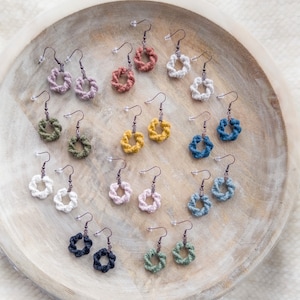 Puede incluir: Una colección de pendientes de macramé hechos a mano en varios colores, incluyendo blanco, rosa, amarillo, azul y verde. Cada pendiente presenta un diseño circular con un gancho de metal. Los pendientes se exhiben en una bandeja de madera.