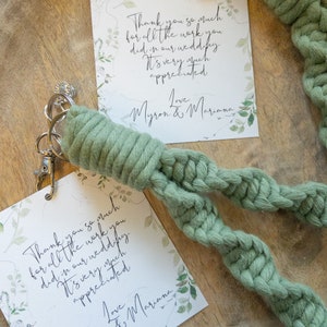 May include: Two thank you cards with green foliage and the text "Thank you so much for all the work you did in our wedding. It's very much appreciated. Love Myron & Mariana" on a wooden surface. A green braided rope keychain with a silver key ring is on top of the cards.