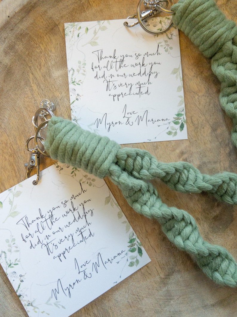 May include: Two thank you cards with green foliage and the text "Thank you so much for all the work you did in our wedding. It's very much appreciated. Love Myron & Mariana" on a wooden surface. A green braided rope keychain with a silver key ring is on top of the cards.