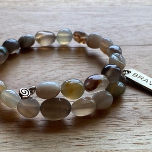 Può includere: Un braccialetto a doppio filo realizzato con perline di agata grigie, marroni e bianche. Il braccialetto ha un ciondolo d'argento con un design a spirale e un'etichetta d'argento con la scritta "BRAVE".