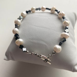 Könnte beinhalten: Ein Armband aus weißen Perlen und schwarzen Edelsteinen mit einem silbernen Verschluss. Das Armband liegt auf einem weißen Kissen.