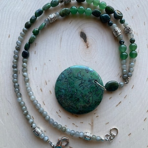 Pode incluir: Um colar artesanal com um pingente grande, redondo e verde com um padrão único. O colar inclui várias contas verdes e cinzentas, espaçadores de metal prateado e um fecho. O colar é exibido em uma superfície de madeira clara.