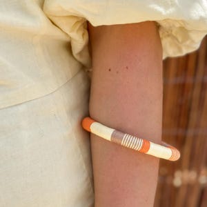 Könnte beinhalten: Nahaufnahme eines geflochtenen Armbands mit orangefarbenen, weißen und braunen Streifen. Das Armband wird am Arm einer Person getragen.