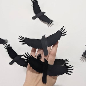 May include: Black paper raven decorations in various stages of flight. The ravens have outstretched wings and are arranged against a white background. The ravens are a dark matte black color.