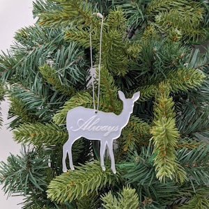 May include: A white, translucent Christmas ornament shaped like a deer with the word "Always" etched on its side. The ornament is hanging on a green artificial Christmas tree.