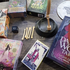May include: A collection of tarot and oracle decks, including "The Herbcrafter's Tarot", "The Rider Tarot", "Celtic Spirit Oracle", "Herbal Astrology Oracle", and "Oracle of the Fairies". There is also a wooden pendulum, a dice that reads "YES", and a set of bones.