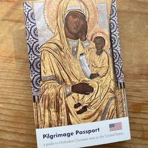 May include: A book titled "Pilgrimage Passport: a guide to Orthodox Christian sites in the United States" with a gold icon of the Virgin Mary and baby Jesus on the cover. The icon is set against a black and white patterned background. The American flag is in the bottom right corner.