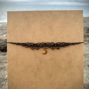 Può includere: Una collana girocollo in macramè marrone scuro con un ciondolo a mezzaluna dorata. La collana presenta piccole perline dorate e colorate. La collana è esposta su una carta marrone su uno sfondo sfocato della spiaggia.