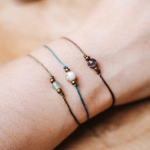 Puede incluir: Tres delicadas pulseras de cuentas en tonos verdes, azules y marrones. Cada pulsera presenta una sola piedra preciosa o cuenta, acentuada con pequeñas cuentas doradas. Las pulseras están ensartadas en finos cordones.